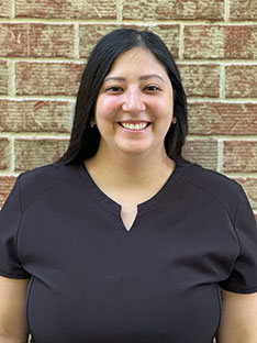 A woman wearing a dark shirt stands against a brick wall, smiling at the camera.