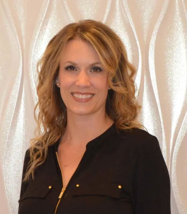 The image shows a woman with blonde hair, wearing a dark top and smiling at the camera.