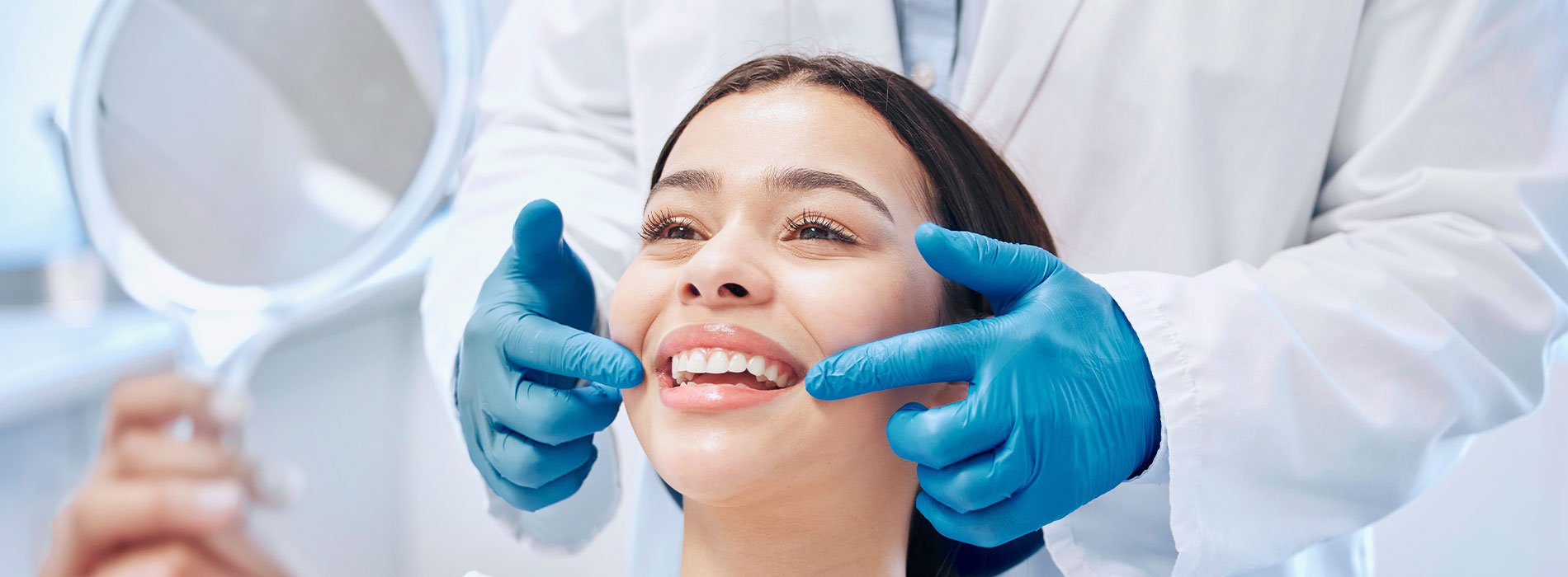 The image shows a person receiving dental care with a dentist using a mirror to inspect their teeth while wearing blue gloves, set against a background of a dental office environment.