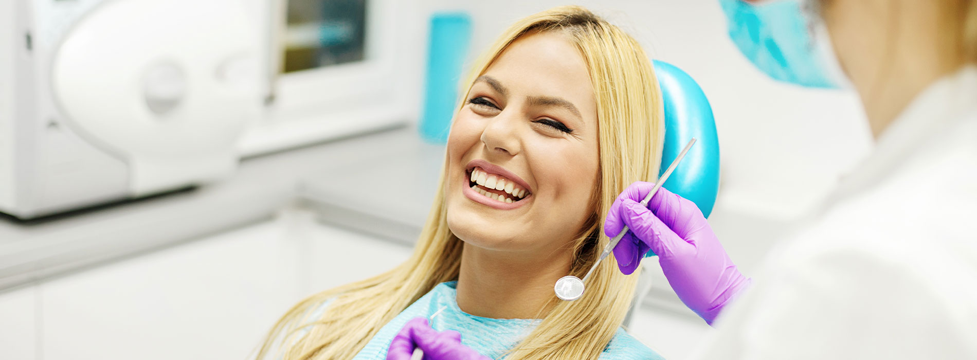 The image shows a woman with blonde hair smiling in a dental chair while receiving oral care from a professional wearing protective gloves and a blue apron, set against the backdrop of a modern dental office.