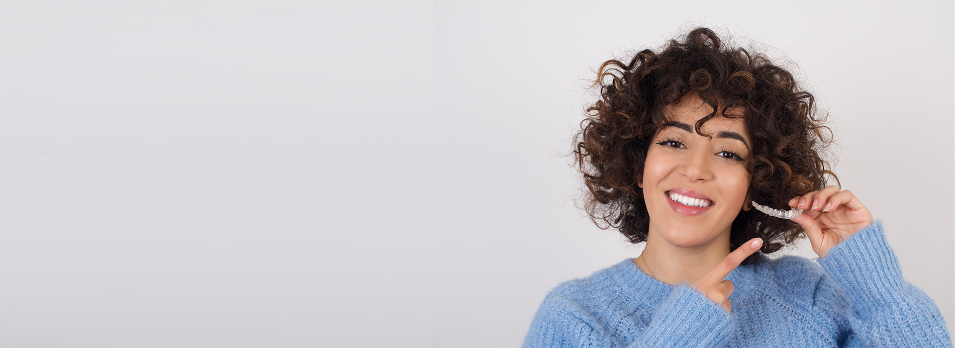 The image shows a person with curly hair smiling at the camera while holding a toothbrush, set against a plain background with a watermark-like design on the right side.