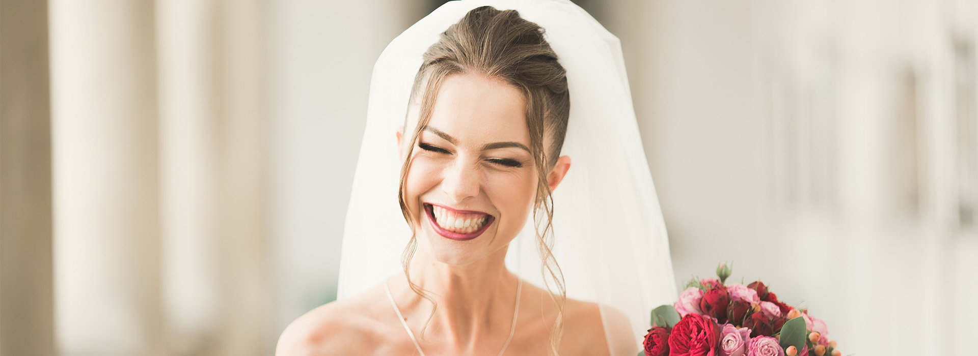 The image shows a woman wearing a white wedding dress and veil, smiling at the camera while holding a bouquet of flowers, with a man standing behind her in a suit with his back to the camera.