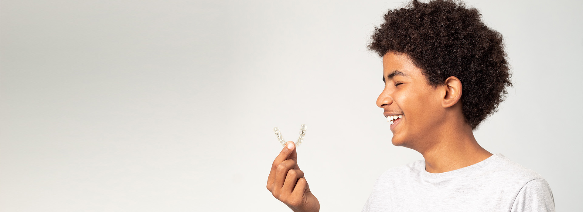The image shows a young person with curly hair smiling at the camera while holding a small object in their hand.