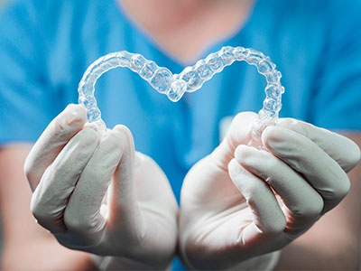 The image shows a person s hands holding a heart-shaped dental retainer made of clear plastic material.