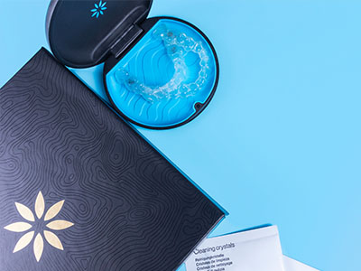 The image shows a blue box with a white label featuring a gold star design, placed on a light blue surface next to a small black container that appears to contain a product, possibly a cream or gel, with a clear lid and a golden spoon resting inside.