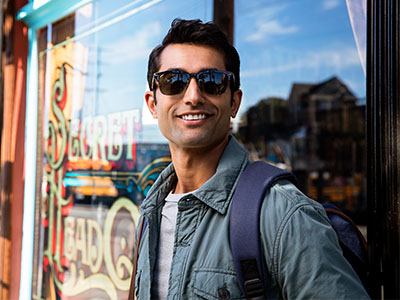 The image shows a man standing outdoors in front of a shop window, wearing sunglasses, a jacket, and carrying a backpack. He has short hair and appears to be looking directly at the camera with a slight smile.
