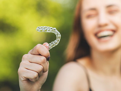The image features a young woman holding up a clear plastic smiley face over her mouth with a joyful expression, set against an outdoor background.