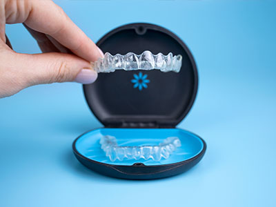 An image showing a hand holding a clear plastic tray containing a set of transparent dental aligners with a blue lid on a tabletop background.