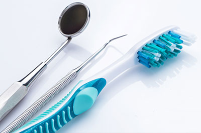 The image shows a collection of dental instruments including a toothbrush with blue bristles and a metal dental pick, laid out on a white surface with a shadow cast beneath them.