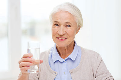 The image depicts an elderly woman holding a glass of water and smiling at the camera.