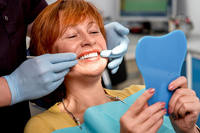 The image depicts a woman sitting in a dental chair with her mouth open, receiving dental care from a professional wearing gloves and a mask. She is holding up a blue toothbrush-shaped object, possibly an oral appliance or dental tool.