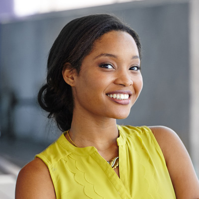 A smiling woman with short hair wearing a yellow top and standing against a backdrop featuring a railing.
