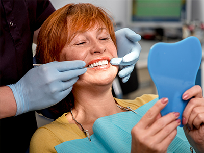 A woman with red hair sits in a dental chair, smiling at the camera while holding up a blue toothbrush, with a dental hygienist standing behind her, preparing for a cleaning session.