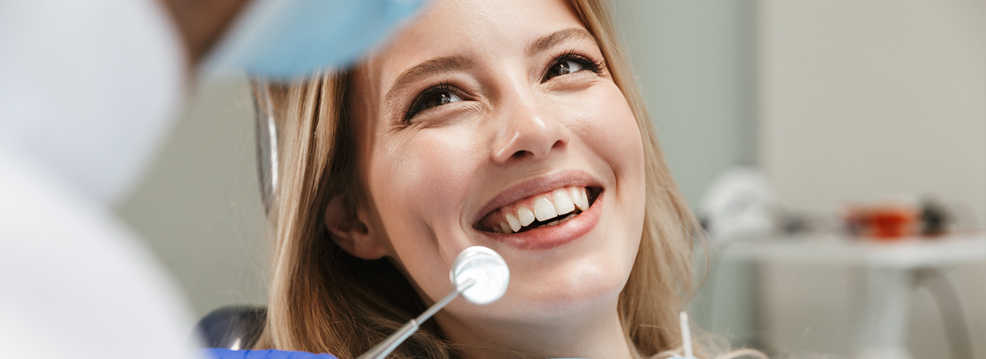 The image shows a young woman with a bright smile, sitting in a dental chair while looking at her reflection in a mirror, with a dental professional standing behind her, wearing protective eyewear and holding an instrument, suggesting she is at a dentist s office for a check-up or procedure.
