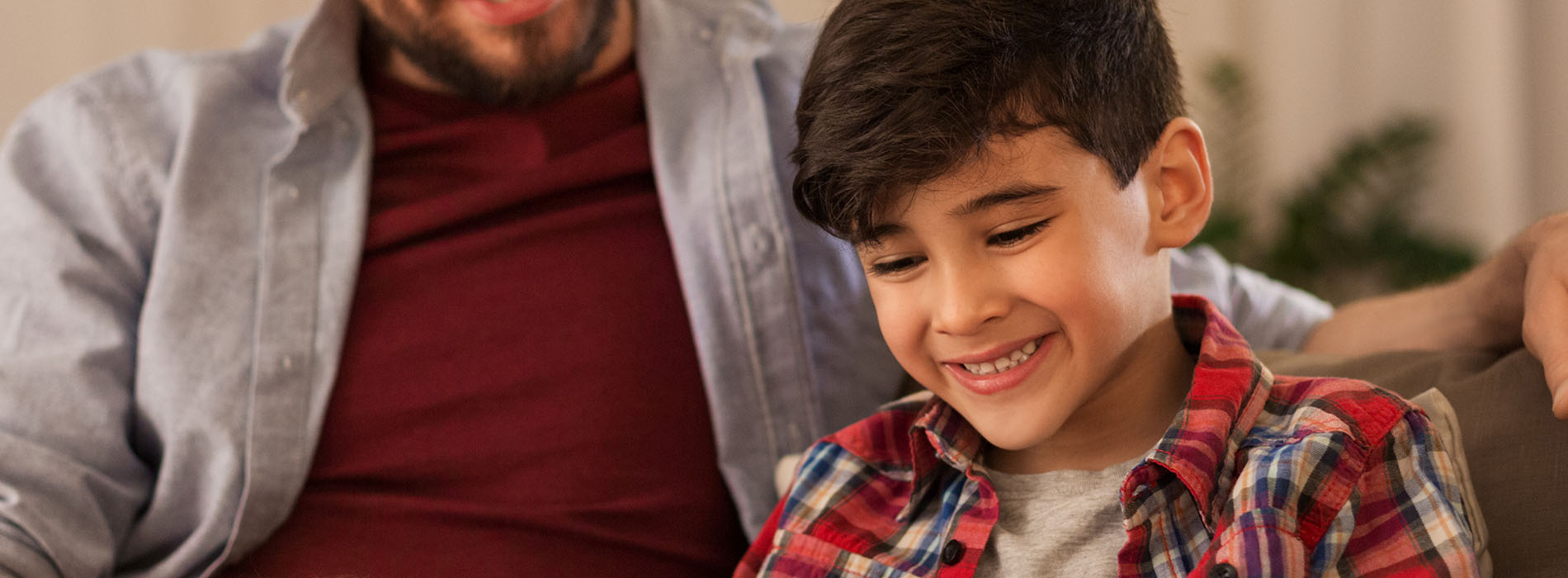 The image shows a young boy sitting on a couch between two adults, smiling at the camera with his arms crossed over his chest.