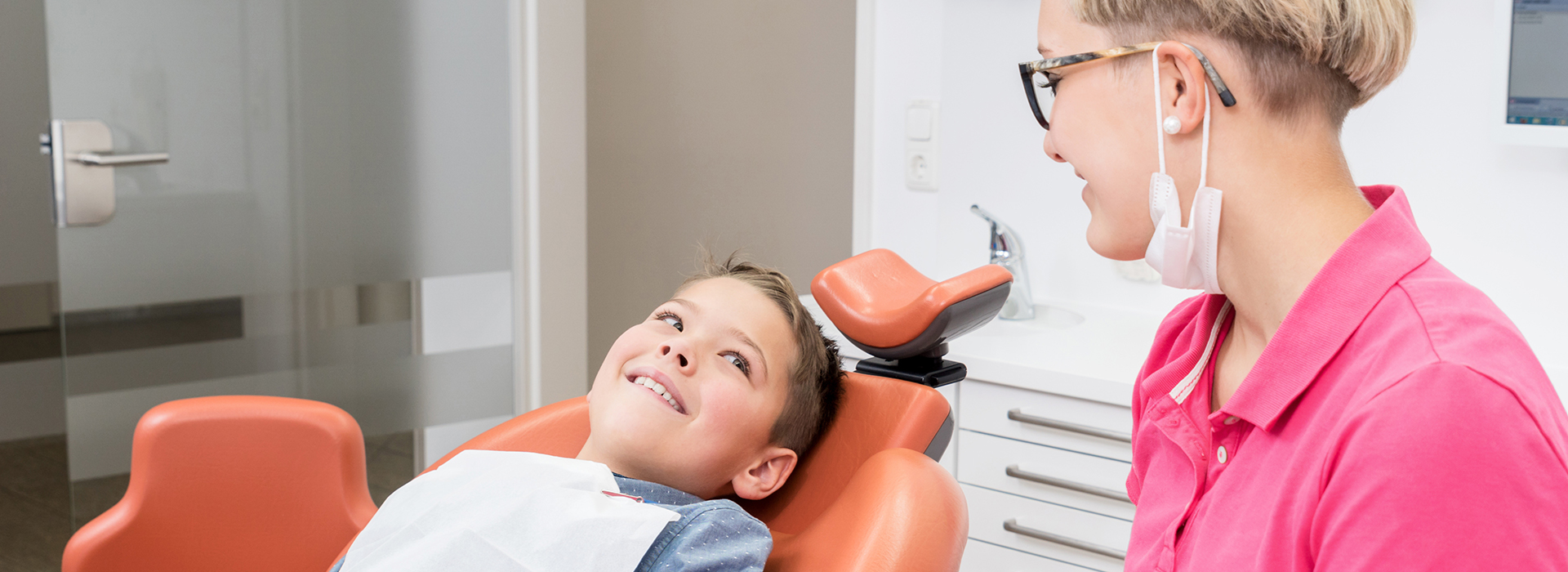 A young boy with glasses sitting in an orange dental chair, smiling at the camera, while a woman in a pink shirt stands behind him, observing his smile.