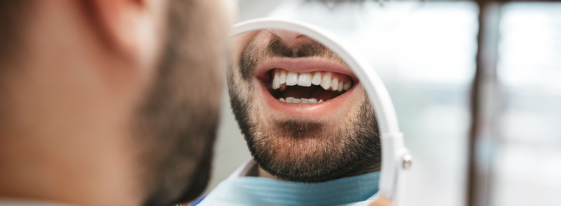 The image features a close-up of a person s face with an open mouth, showing teeth, set against a blurred background that includes another person partially visible on the right side, suggesting a dental or medical setting.