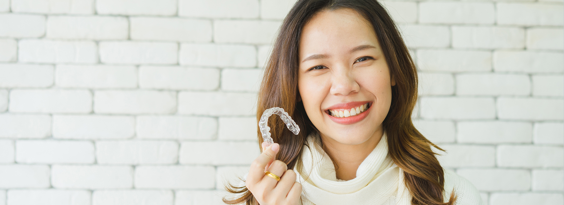 A woman holding up a toothbrush with a smile, set against a brick wall background.