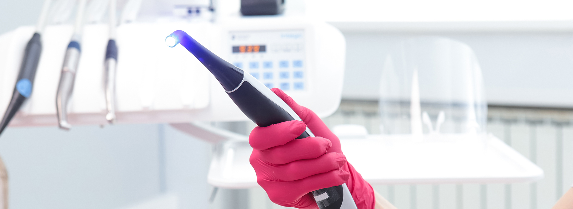 The image shows a person wearing red gloves holding a blue electric toothbrush, set against a backdrop with various medical equipment and a white countertop.
