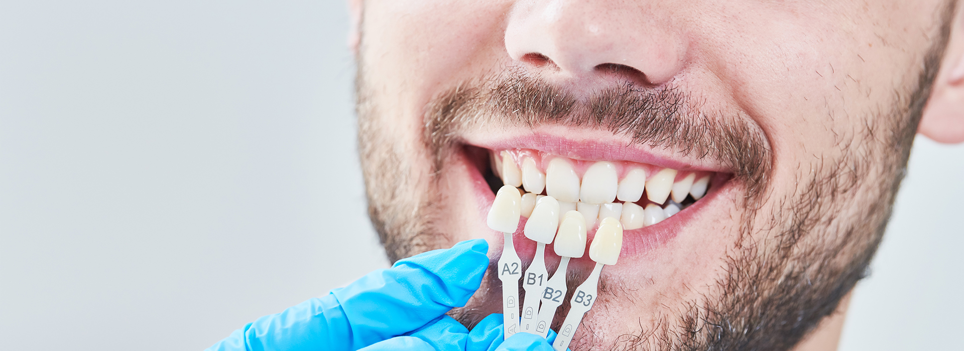 A man with a smiling expression is shown holding a toothbrush to his teeth while wearing blue gloves and looking directly at the camera.