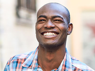 A smiling man with short hair, wearing a plaid shirt, stands outdoors against a backdrop of an urban street scene.