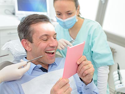The image shows a man sitting in a dental chair with a cardboard sign on his chest, smiling at the camera while holding up the sign, which has text on it. Behind him, there are medical professionals, including a dentist and a nurse, who appear to be engaged in a discussion or examination of the man s teeth. The setting suggests a dental office environment with various dental equipment visible.