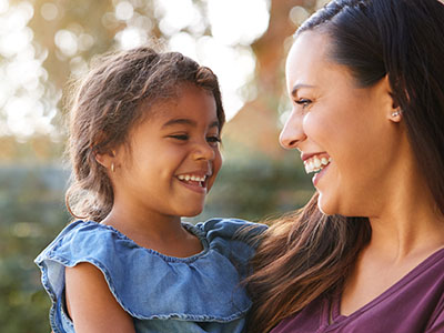 A woman and a young child are smiling at each other outdoors.