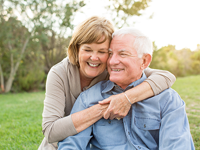 A man and woman are sitting closely together outdoors, embracing each other with smiles on their faces.