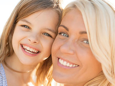The image shows two individuals, an adult woman with blonde hair and a young girl with dark hair, sharing a joyful moment together with smiles on their faces. They are outdoors during daylight, and both appear happy and content.