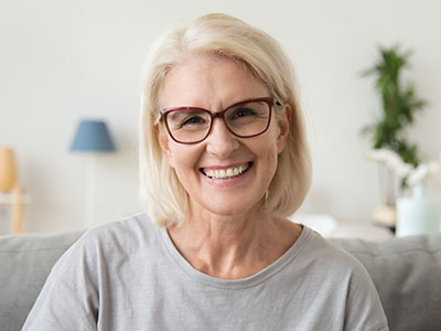 The image shows a woman with short blonde hair smiling at the camera, wearing glasses, a light-colored top, and seated indoors.