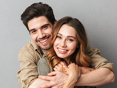 A young couple hugging closely, both smiling, with a man wearing a brown shirt and a woman in a light-colored top.