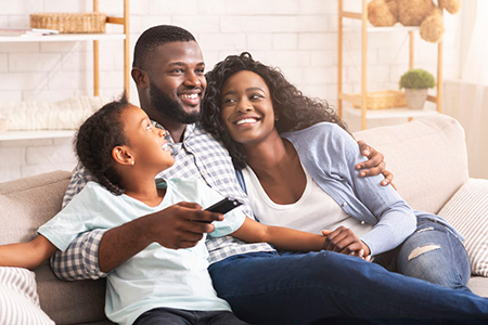 A family of four sitting together on a couch, with a man holding a child, smiling at the camera.