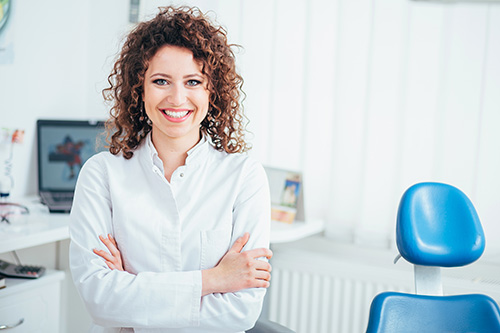 The image features a woman with curly hair smiling at the camera, dressed in a white shirt and standing in an office setting with dental equipment visible in the background.