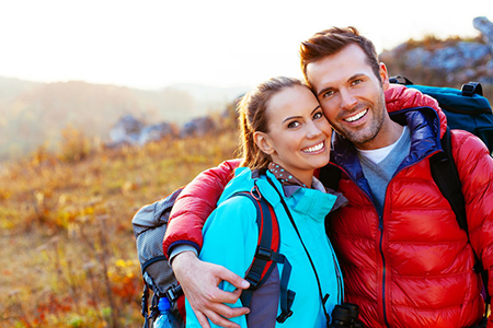 The image shows a man and woman posing together outdoors during daylight, with the man wearing a backpack and both dressed in casual outdoor attire.