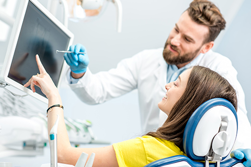The image shows a dental professional assisting a patient during a dental appointment, with the professional standing behind the patient who is seated in a dental chair, pointing at something on a computer screen.