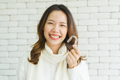 The image features a woman holding a smiley face emoji with her right hand, smiling at the camera, against a brick wall background.