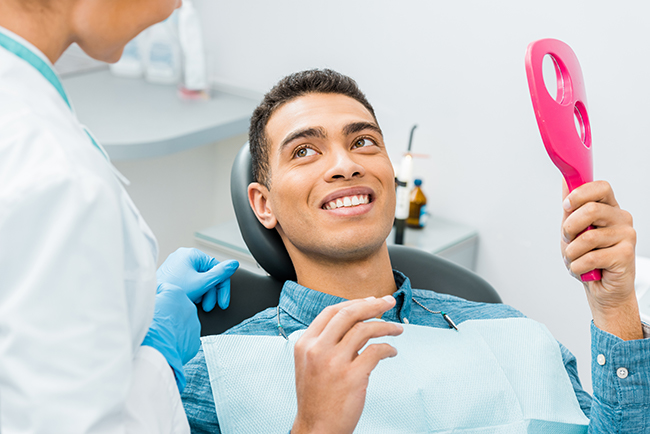The image depicts a person sitting in a dental chair with a smile, holding up a pink object towards their face, while being attended by a dental professional.