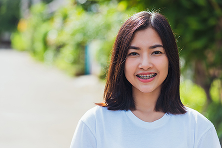 The image shows a young woman with short hair smiling at the camera. She has a light complexion, wears a white top, and stands outdoors against a backdrop of greenery and a clear sky.