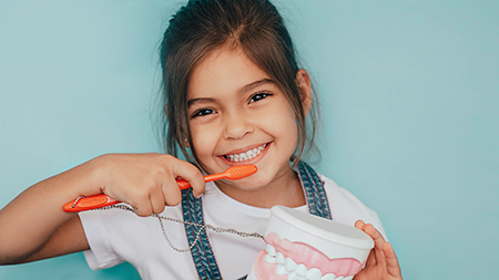 A young girl brushing her teeth with a red toothbrush, smiling at the camera.