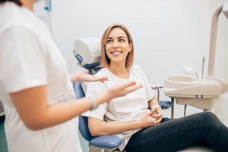 A woman sitting in a dental chair with her hands clasped together, smiling at someone outside the frame, while being attended by dental professionals.