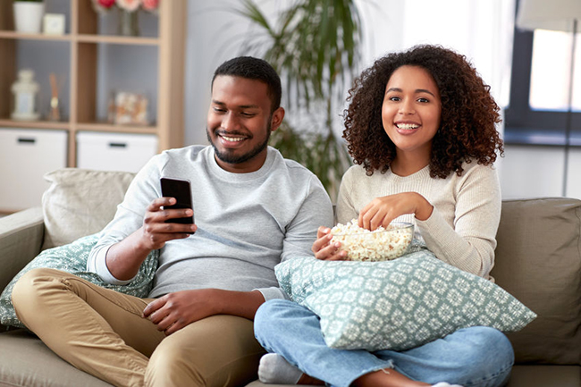 A man and woman sitting on a couch, sharing a moment together while enjoying popcorn, with both individuals smiling and seemingly engaged in watching something on a screen out of frame.