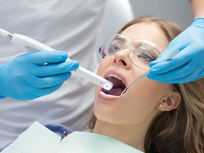 A woman receiving dental treatment with an electric toothbrush held by a dental professional wearing protective gloves and a face mask, set against a clinical background.