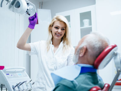 A dentist attending to an elderly patient in a dental chair, with a smiling young woman standing behind them holding a small device.