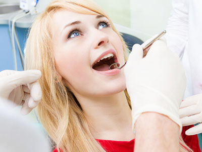 The image shows a woman sitting in a dental chair with her mouth open wide, receiving dental treatment. A dentist is working on her teeth.