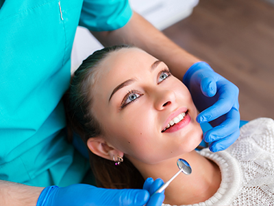 The image shows a woman sitting in a dental chair with her eyes closed, receiving dental care from a professional wearing a surgical mask and gloves.