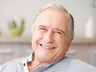 A man with white hair and glasses, smiling and leaning back in a relaxed posture, appears to be in his senior years, wearing a blue shirt and sitting in front of a kitchen counter.
