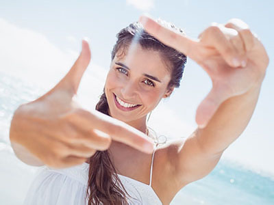 A woman is holding up two fingers with a smile on her face against a bright beach background.